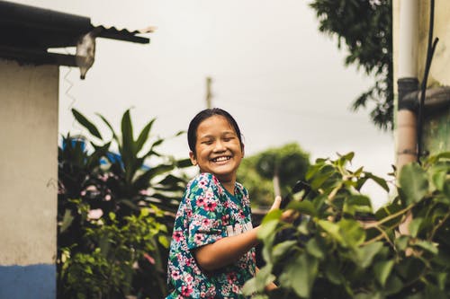 闭着眼睛站在绿叶植物旁边的小女孩微笑着的照片 · 免费素材图片