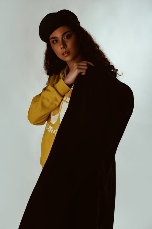 黄色连帽衫，黑色贝雷帽和黑色外套摆在白色背景前的女人的照片 · 免费素材图片