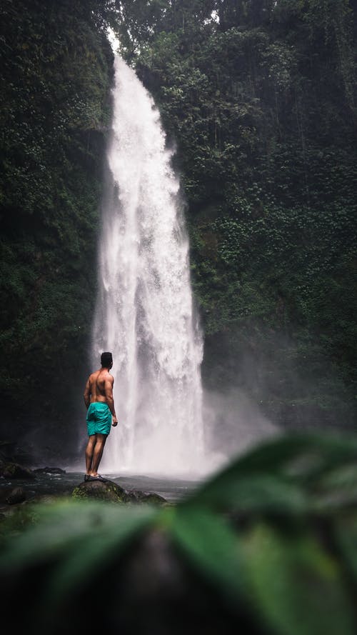 站在瀑布附近的岩石上的裸照男子的照片 · 免费素材图片