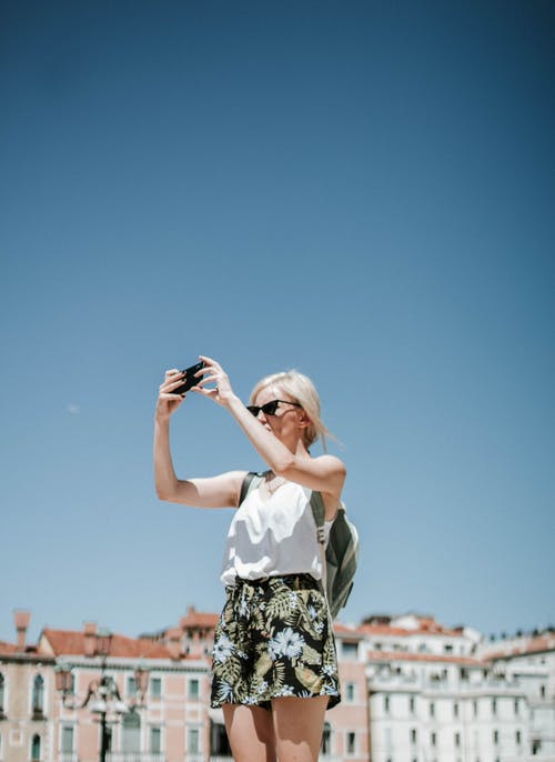 女人穿着黑色和白色的花卉短和白色背心的照片使用黑色的智能手机拍照 · 免费素材图片