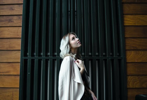 女人穿着白色外套站在黑色金属门前仰望的侧视图照片 · 免费素材图片