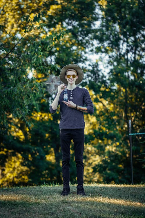 微笑的男人，在棕色的帽子和太阳镜举起一个玻璃瓶的照片 · 免费素材图片