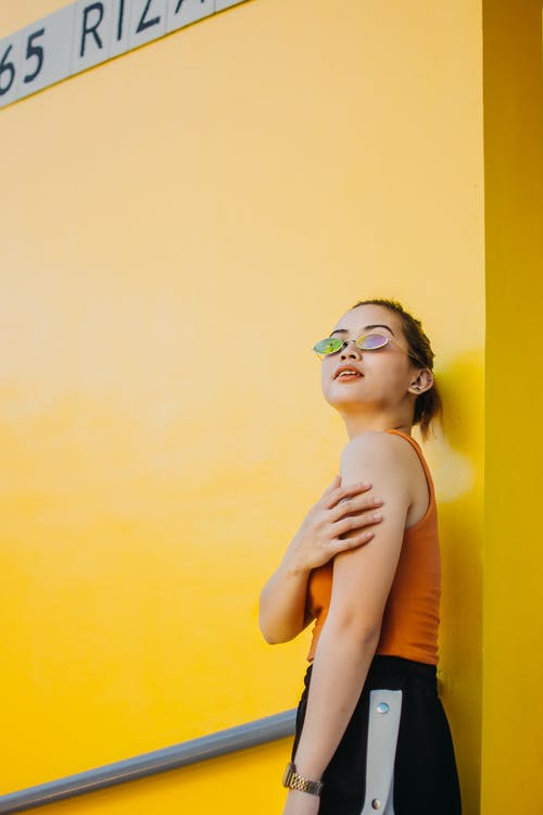 戴墨镜，橙色上衣和黑色下装站在黄墙旁边的女人的照片 · 免费素材图片