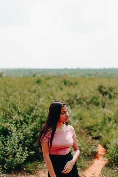 选择性聚焦照片女人粉红色的露肩上衣和黑色的裙子看着别处与绿色的植物在背景中 · 免费素材图片