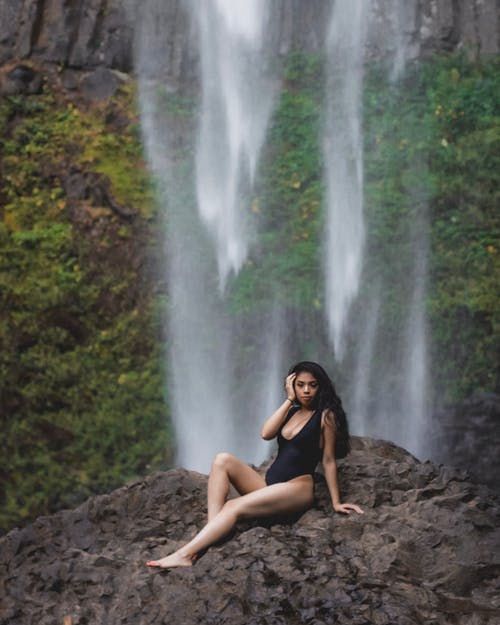 摆在岩石上与瀑布在背景中的黑色泳装的女人的照片 · 免费素材图片