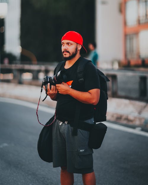 戴着黑色t恤和红色帽子拿着照相机的人 · 免费素材图片