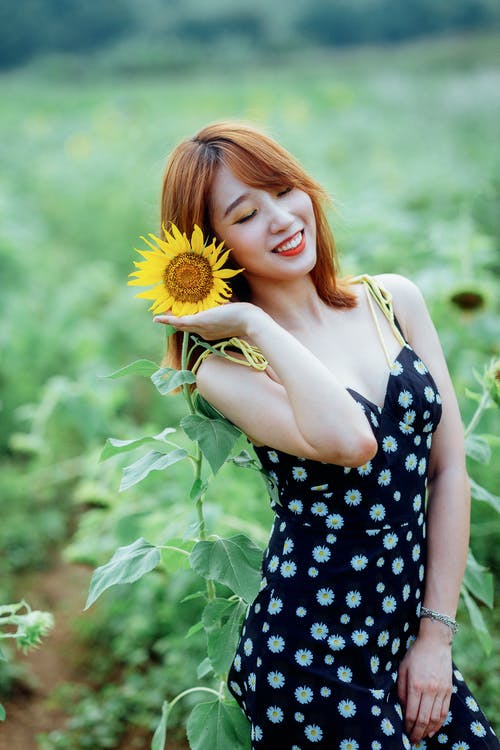 向日葵在她旁边闭着眼睛摆姿势的黑色花裙子的微笑妇女的选择性聚焦照片 · 免费素材图片