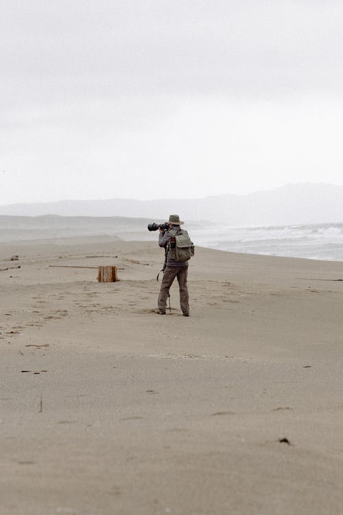 男子手持相机站在海边 · 免费素材图片
