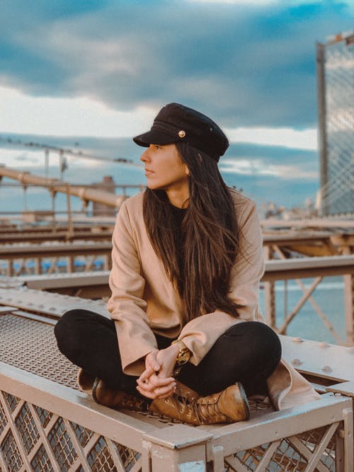 女人穿着棕色长袖衬衫和黑色的帽子，看着别处时坐在金属表面上的选择性焦点照片 · 免费素材图片