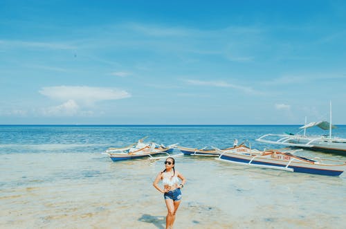 女人穿着白色比基尼上衣和蓝色短裤与两手叉腰站在蓝色和白色的天空下海附近 · 免费素材图片