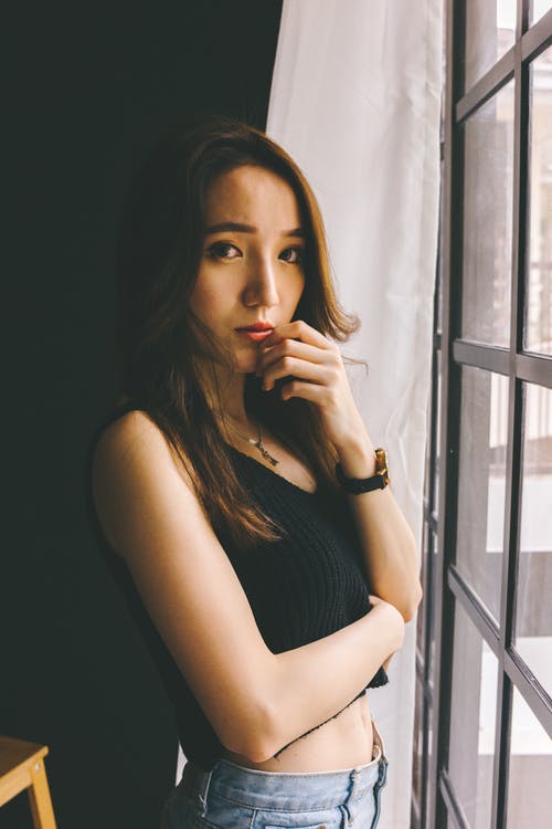 黑色无袖作物上衣和蓝色牛仔裤摆在窗户旁的女人的照片 · 免费素材图片