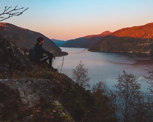 男子坐在悬崖上面临着平静的水体 · 免费素材图片