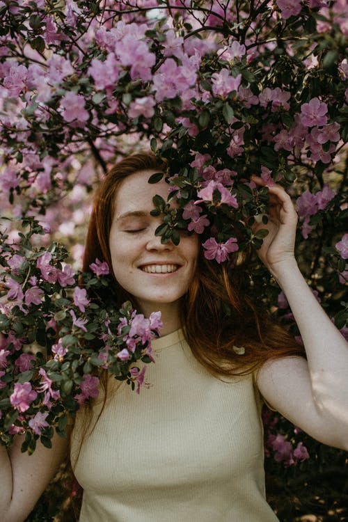 闭着眼睛摆在粉红色的开花植物下的微笑妇女的照片 · 免费素材图片