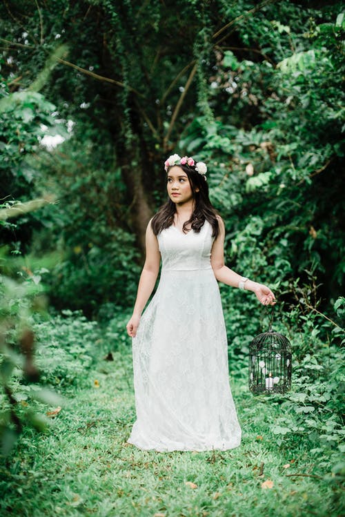 女人穿着白色礼服和花冠拿着鸟笼烛台站在树林中间的照片 · 免费素材图片