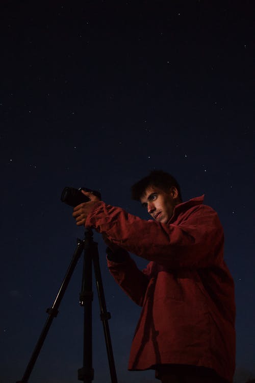 穿着红色夹克的人站立在三脚架附近的照相机在夜间 · 免费素材图片