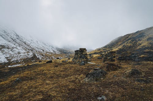 低角度拍摄的成堆的岩石在枯萎的土地上，在白雪皑皑的群山与浓密的云朵旁边 · 免费素材图片