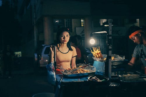 女人在晚上在棍子上吃街头食品 · 免费素材图片