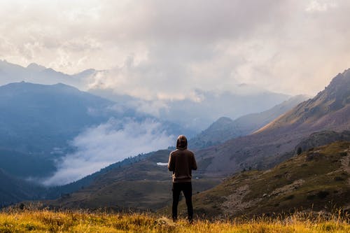 穿着棕色套头衫和黑色牛仔裤的人站在山顶上，风景秀丽 · 免费素材图片