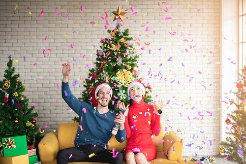 男人和女人戴着圣诞老人的帽子，坐在沙发上弹出包围圣诞树的五彩纸屑 · 免费素材图片