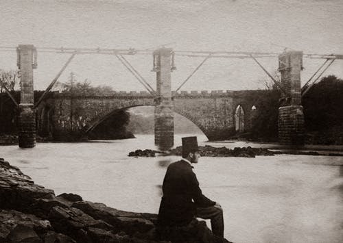 戴着帽子和西装坐在桥边河边的灰度照片男子 · 免费素材图片