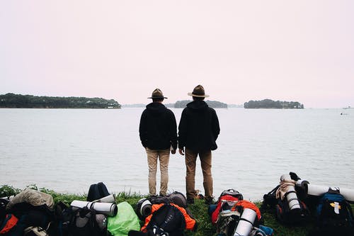 两名戴着帽子和黑色夹克的男人站在水体附近 · 免费素材图片