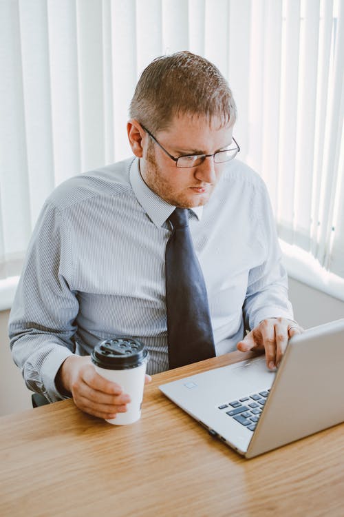 拿着咖啡杯和使用便携式计算机的灰色衬衫的男人 · 免费素材图片