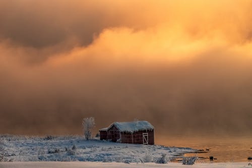 靠近河的棕色谷仓在多云的天空和冬天的白雪覆盖的地面下 · 免费素材图片