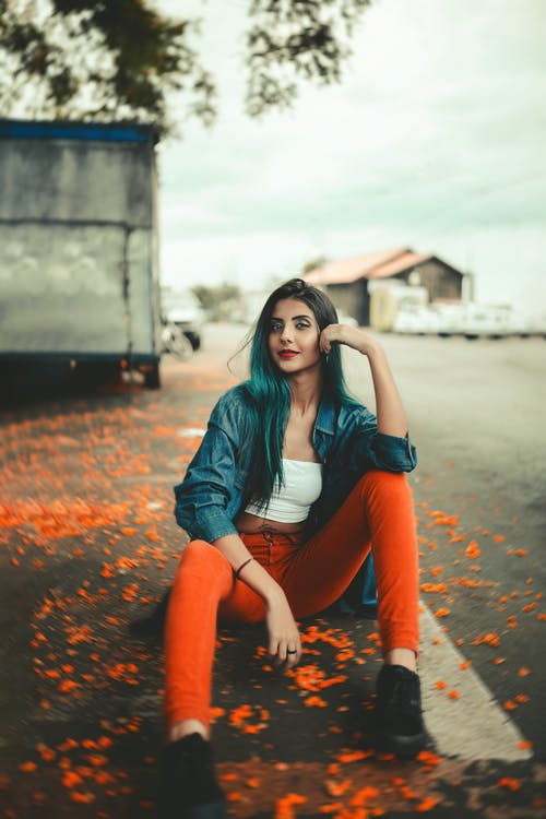 蓝色牛仔夹克和橙色的裤子，坐在马路上摆姿势的女人的照片 · 免费素材图片