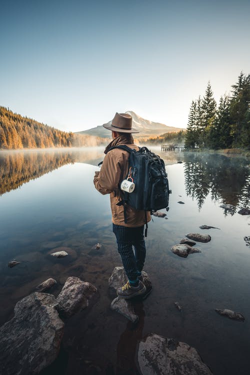 棕色夹克和棕色帽子站在湖附近的岩石上的人 · 免费素材图片