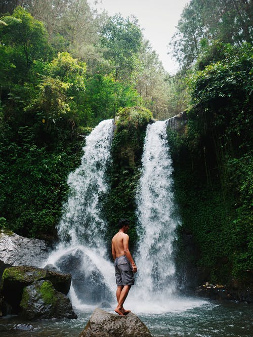 男子站在瀑布前的岩石上 · 免费素材图片