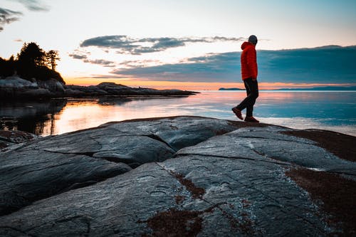 穿红色夹克的男人站在石头上看着水的身体 · 免费素材图片