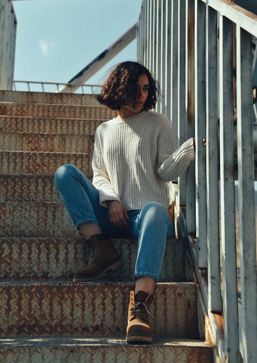 女人穿着白色的毛衣和蓝色牛仔裤，坐在楼梯上 · 免费素材图片