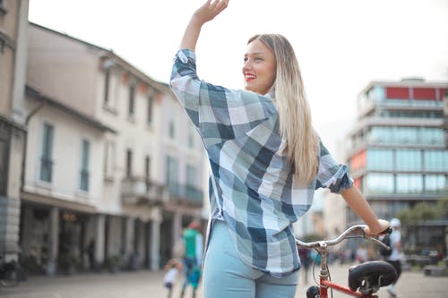 后视图的微笑的女人，穿花格子衬衫和蓝色牛仔牛仔裤，站着自行车挥舞着的照片 · 免费素材图片