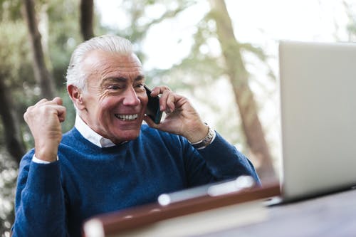兴奋的老人，穿着蓝色毛衣坐在桌旁，一边使用笔记本电脑在电话上交谈的选择性聚焦照片 · 免费素材图片