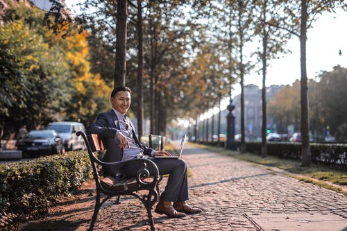 坐在公园的长凳上，使用笔记本电脑的灰色西装的男人 · 免费素材图片
