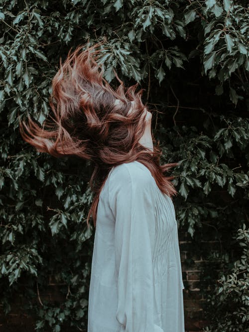 女人穿着白色长袖连衣裙站在附近绿叶植物做头发翻转的侧视图照片 · 免费素材图片