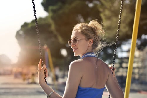 微笑的女人，在蓝色上衣和太阳镜坐在秋千上回望的选择性焦点照片 · 免费素材图片
