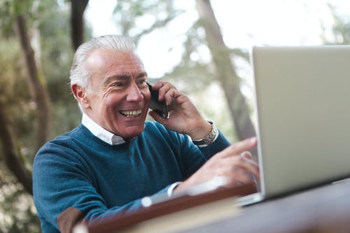 微笑的老人穿着蓝色毛衣，坐在桌旁，一边使用笔记本电脑在电话上交谈的选择性焦点照片 · 免费素材图片