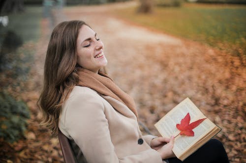 笑的女人，穿着棕色大衣，坐在公园的长椅上拿着一本书的选择性焦点照片 · 免费素材图片
