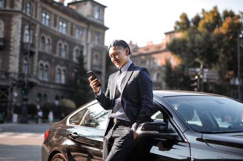 穿着黑色西装的男人，靠在黑色汽车旁边时使用手机的选择性聚焦照片 · 免费素材图片