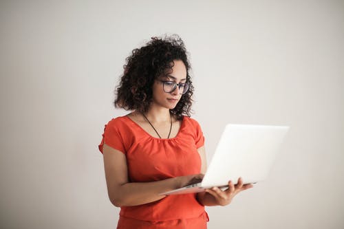站立时使用一台笔记本电脑的橙色上衣的女人的照片 · 免费素材图片