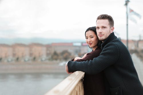 站在栏杆从背后拥抱棕色外套的女人的男人的选择性聚焦照片 · 免费素材图片