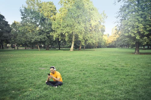 黄色的t恤和黑色的裤子，坐在绿色的草地上，一边听耳机上的音乐，一边使用他的电话的人的照片 · 免费素材图片