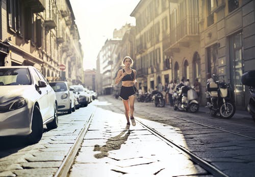 女人在慢跑在铺好的街道上的耳机听音乐的照片 · 免费素材图片