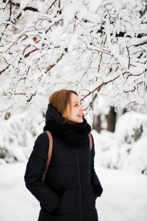 黑色冬季大衣站在雪覆盖树附近微笑的女人的照片 · 免费素材图片