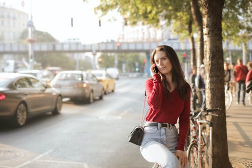 女人穿着红色长袖毛衣和蓝色牛仔裤坐在电话上交谈时坐在金属栏杆上的选择性焦点照片 · 免费素材图片