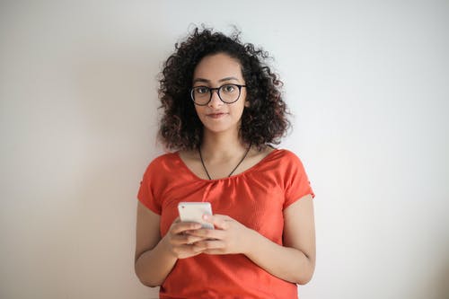 红顶和黑色镜框的眼镜，拿着白色的智能手机的女人的照片 · 免费素材图片