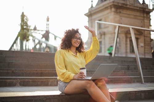 微笑的女人，穿着黄色长袖衬衫和灰色的短裤，挥舞着坐在笔记本电脑上的膝上，拿着一杯咖啡坐在楼梯上的选择性焦点照片 · 免费素材图片
