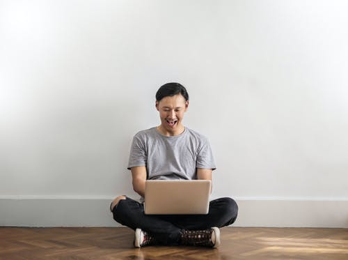 使用笔记本电脑时坐在木地板上坐在灰色的t恤和黑色牛仔裤的男人笑的照片 · 免费素材图片