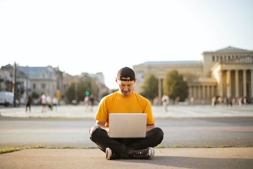 使用他的笔记本电脑坐在地上的黄色t恤和黑色牛仔裤的男人的选择性聚焦照片 · 免费素材图片
